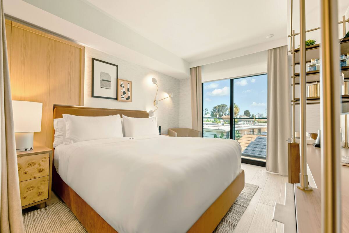 A hotel room with a large bed, a nightstand, a desk, a chair, a bookshelf, and a large window with a view.