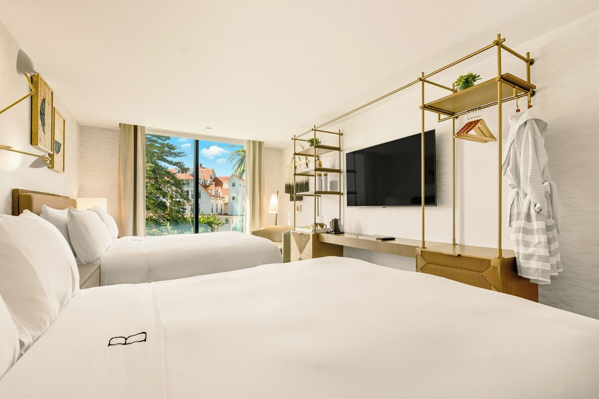 A hotel room with two beds, a TV, a desk, and a balcony with a view of buildings.