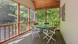 A covered outdoor area with a wooden deck, a glass table, and chairs, overlooking a forested area. -  Lake Kanasatka - Private beach access. Your perfect vacation starts right here! (Moultonborough)