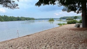 A sandy beach with a calm lake, trees, and a small island. -  Lake Kanasatka - Private beach access. Your perfect vacation starts right here! (Moultonborough)