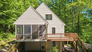 A two-story cabin with a balcony and a wooden deck, surrounded by trees. -  Lake Kanasatka - Private beach access. Your perfect vacation starts right here! (Moultonborough)