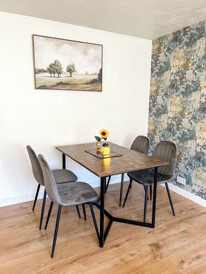 Enjoy meals in this stylish dining area with a wooden table and four unique chairs, a vase with a sunflower, and a framed painting.