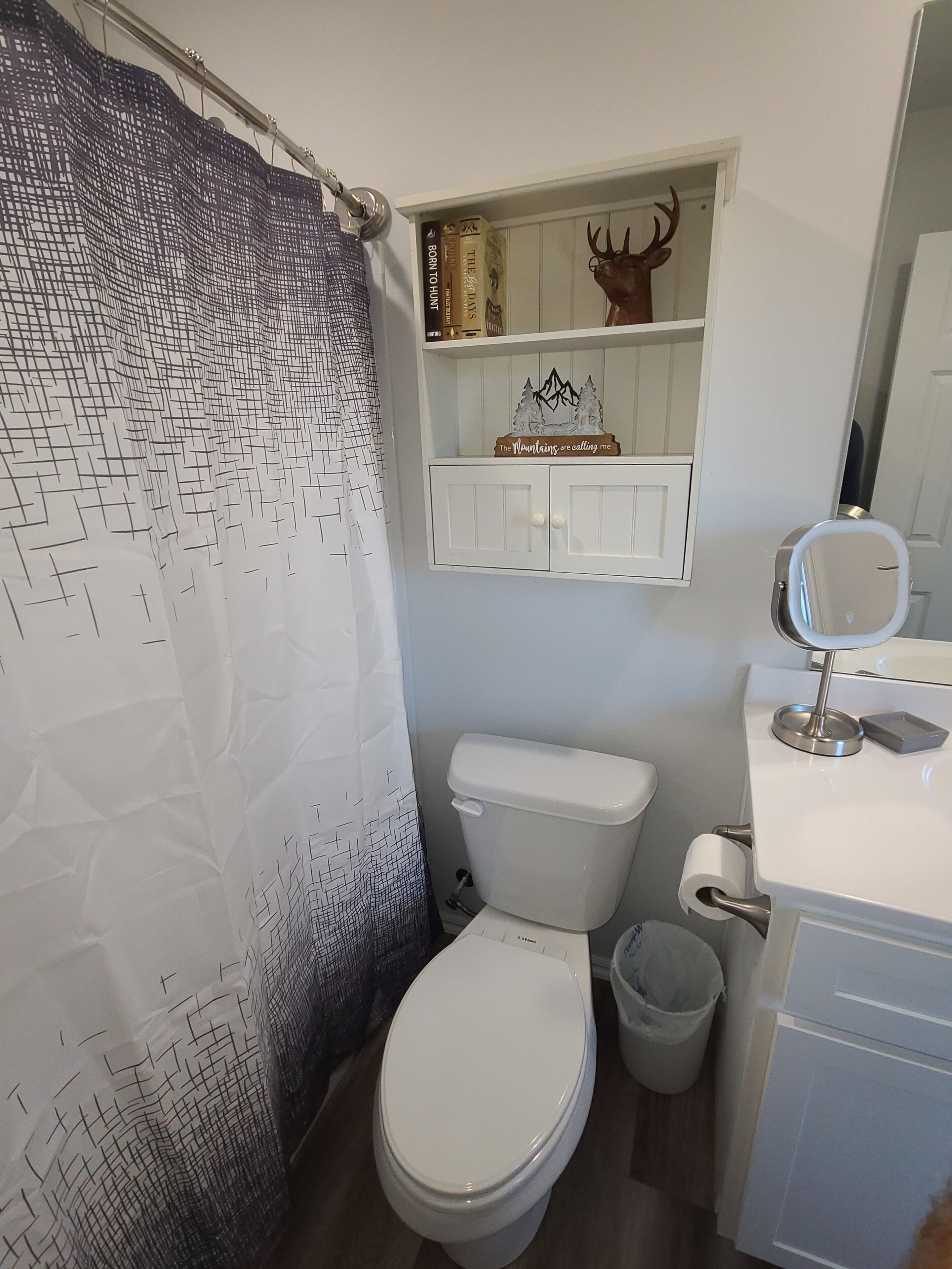 Een badkamer met een wit toilet, een witte wastafel met spiegel, een plank met boeken en een decoratie van een hertenhoofd, en een douchegordijn met een geometrisch patroon.