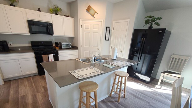 A modern kitchen with white cabinets, a central island with stools, a black refrigerator, and a microwave. - Mac's Cozy Farmhouse (Stillwater)