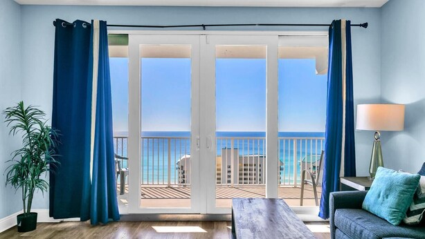 A room with a wooden floor, a sofa, a table, a lamp, and a view of the ocean through a sliding glass door with blue curtains. - Laketown Wharf 1608- Turtle Tower (Panama City Beach)