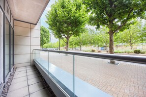 Un bâtiment moderne doté d’un balcon en verre donnant sur une zone pavée agrémentée d’arbres et bordée d’une route.