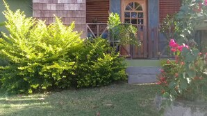 Un edificio a due piani con facciata in pietra, un balcone in legno e un piccolo giardino con arbusti verdi e fiori rosa.