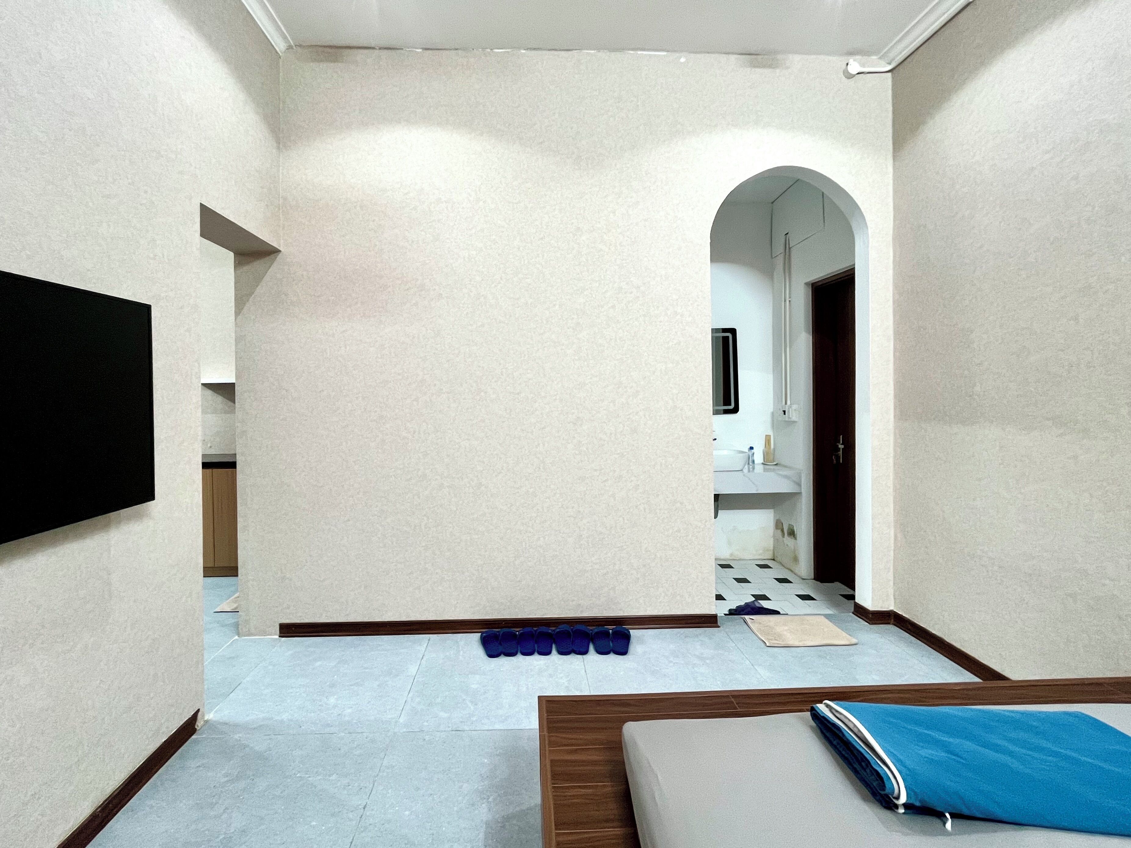 Habitación de hotel con cama, televisor montado en la pared, baño con lavamanos y espejo, y pasillo con piso de baldosas.