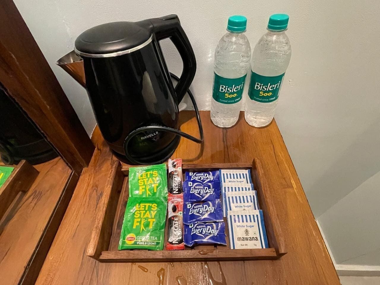 Ein Tablett mit einer schwarzen Kaffeemaschine, zwei Bisleri-Wasserflaschen, grünen und blauen Teebeuteln sowie weißen Zuckerportionen.