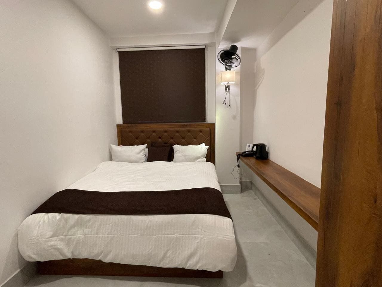 Een net opgemaakt bed met een bruin hoofdeinde, een nachtkastje met een lamp, en een ventilator bevestigd aan de muur.