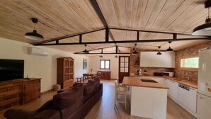 A spacious living area with a wooden ceiling, a brown sofa, a dining table, and a kitchen with white cabinets and a wooden countertop.