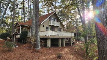 A two-story wooden cabin with a balcony, surrounded by trees and a forest floor covered with pine needles.