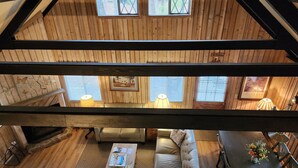 Une pièce de style « loft », avec des poutres apparentes, un canapé, une table basse et une table à manger avec des chaises. Il y a deux fenêtres équipées de stores, une lampe et un tableau encadré accroché au mur.