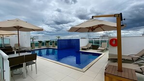 An indoor swimming pool with two umbrellas, lounge chairs, and a view of the sea under a cloudy sky. - OZPED Flats Particulares Biarritz (Brasília)