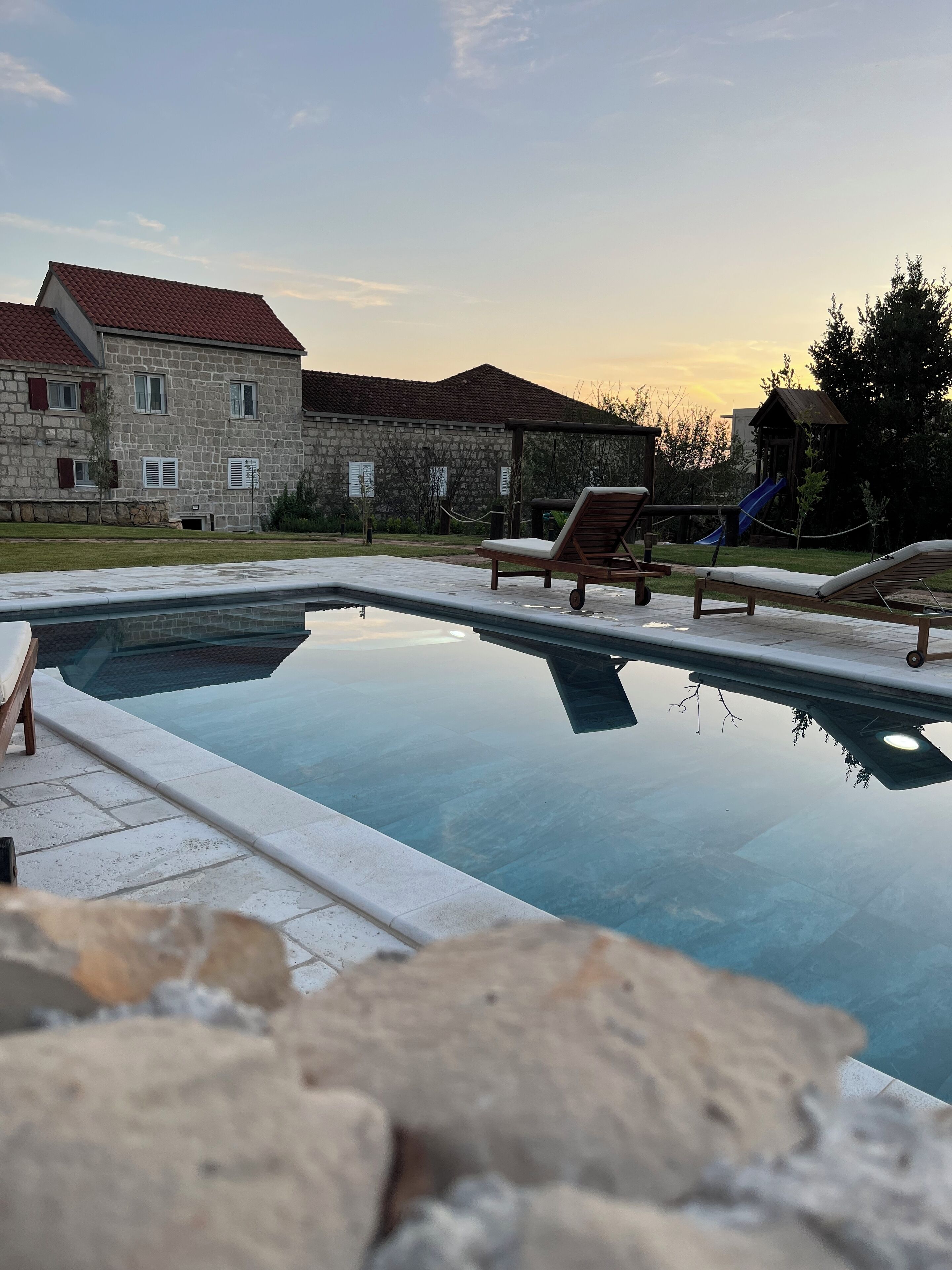 Ein Schwimmbecken mit Liegestühlen, ein Steinhaus und ein Spielplatz im Hintergrund.