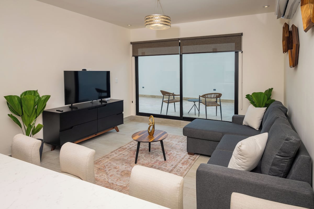 Een moderne woonkamer met een flatscreen televisie, een donkergrijze bank, een salontafel en een grote schuifpui naar een balkon.