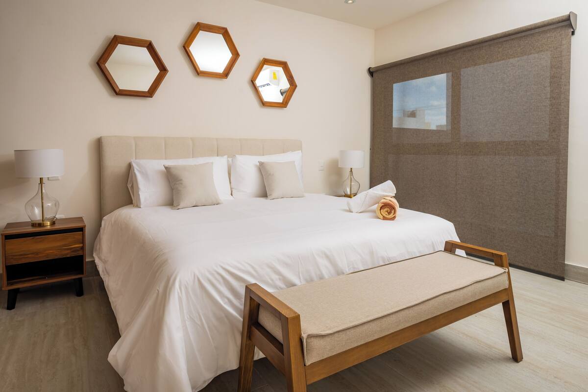 Een keurig opgemaakt bed met een houten bankje, twee nachtkastjes met lampen, en drie zeshoekige spiegels aan de muur.