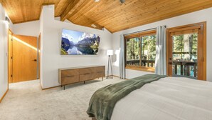 Un dormitorio con una cama grande, un ventilador de techo, un televisor montado, un aparador de madera y un ventanal con vistas a árboles.
