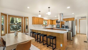 Una cocina moderna con armarios de madera, una isla central con taburetes, electrodomésticos de acero inoxidable y ventanas amplias con vistas a árboles.