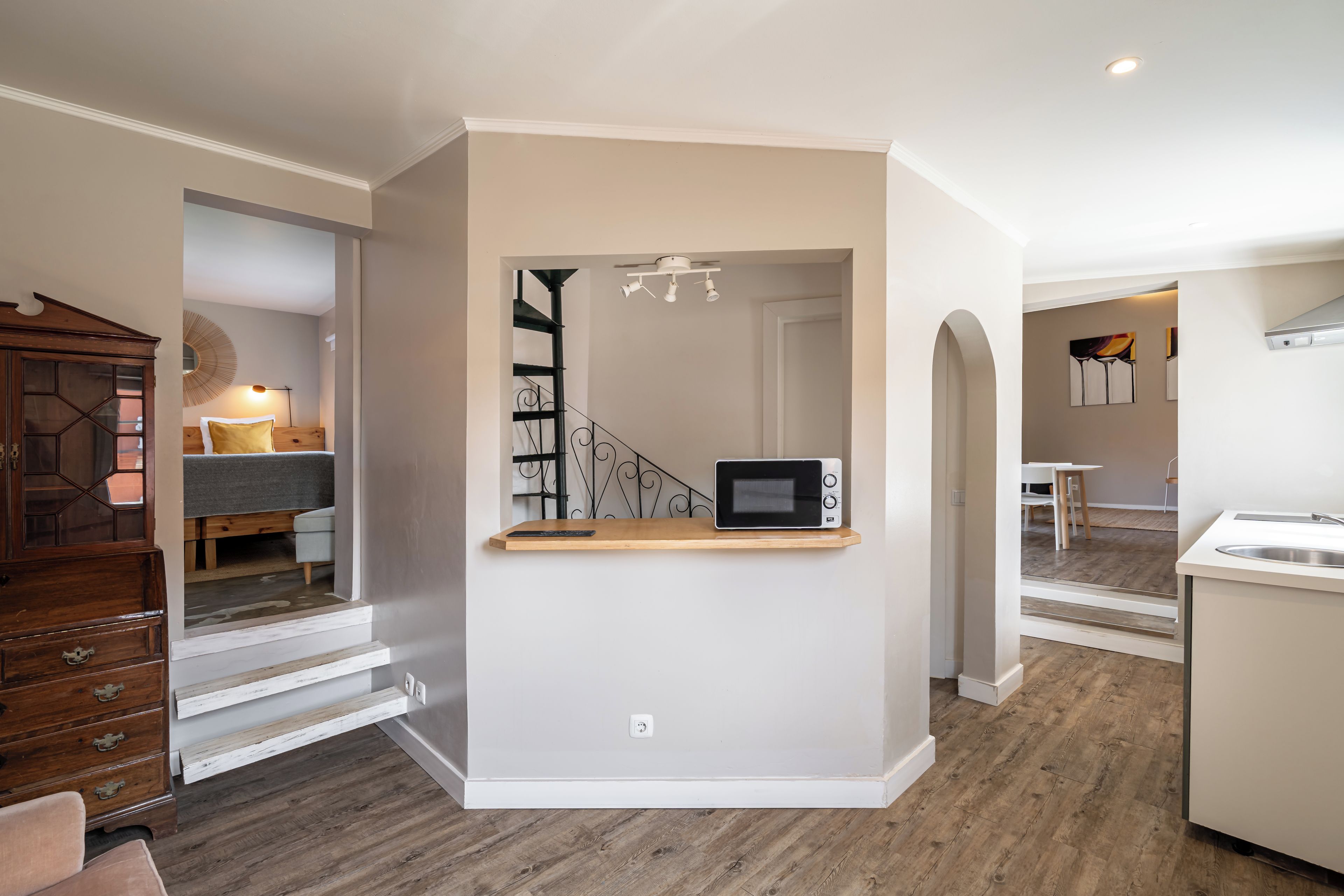Cozinha compacta com micro-ondas em prateleira de madeira, escada que leva a um quarto e área de jantar com mesa e cadeiras brancas.