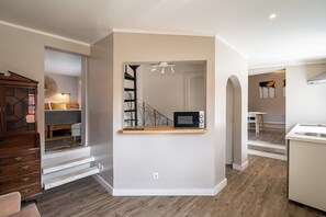 Een kleine keuken met een magnetron op een houten plank, een trap naar een slaapkamer, en een eethoek met een witte tafel en stoelen.