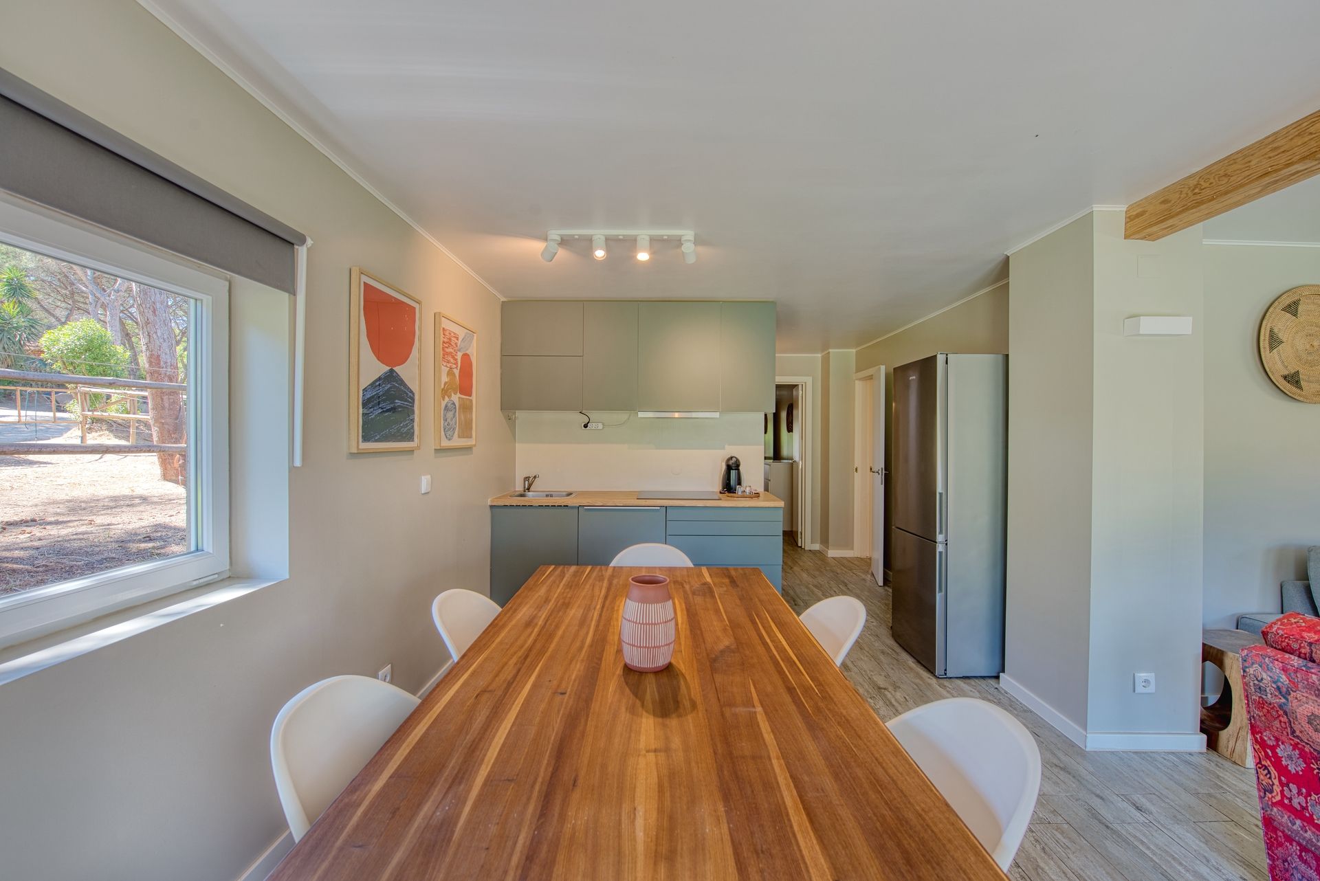 Un comedor con mesa de madera, sillas blancas, una cocina al fondo y vista al exterior.