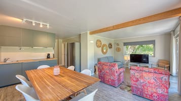 A modern kitchen with a wooden dining table and white chairs, a living area with patterned sofas, a flat-screen TV, and a large window with a view of greenery.
