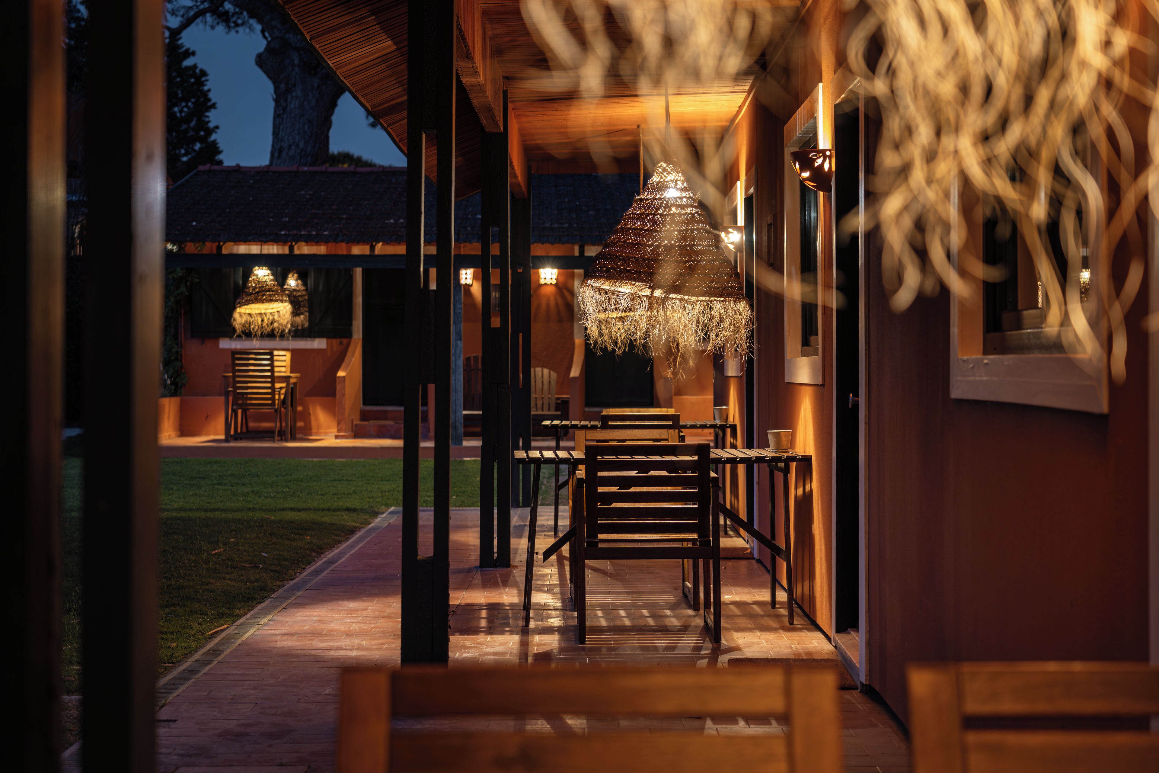 Un área de comedor al aire libre con mesas y sillas de madera, un paraguas de paja con luz y vista a un edificio al fondo.