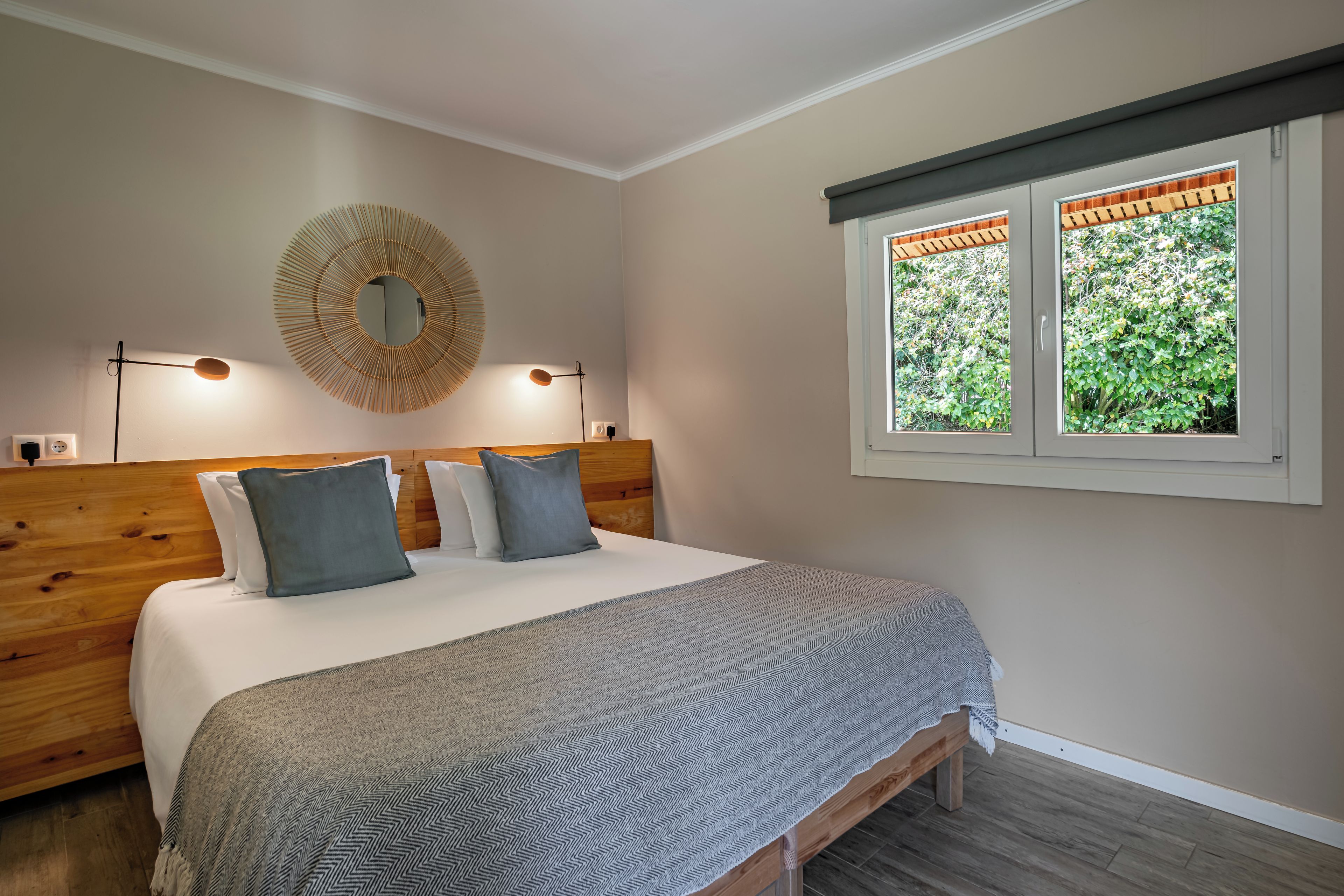 A bedroom with a bed, two bedside lamps, a wall-mounted mirror, and a window with a view of greenery.