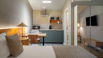 A compact hotel room with a bed, a desk, a chair, a kitchenette, a TV, and a window.