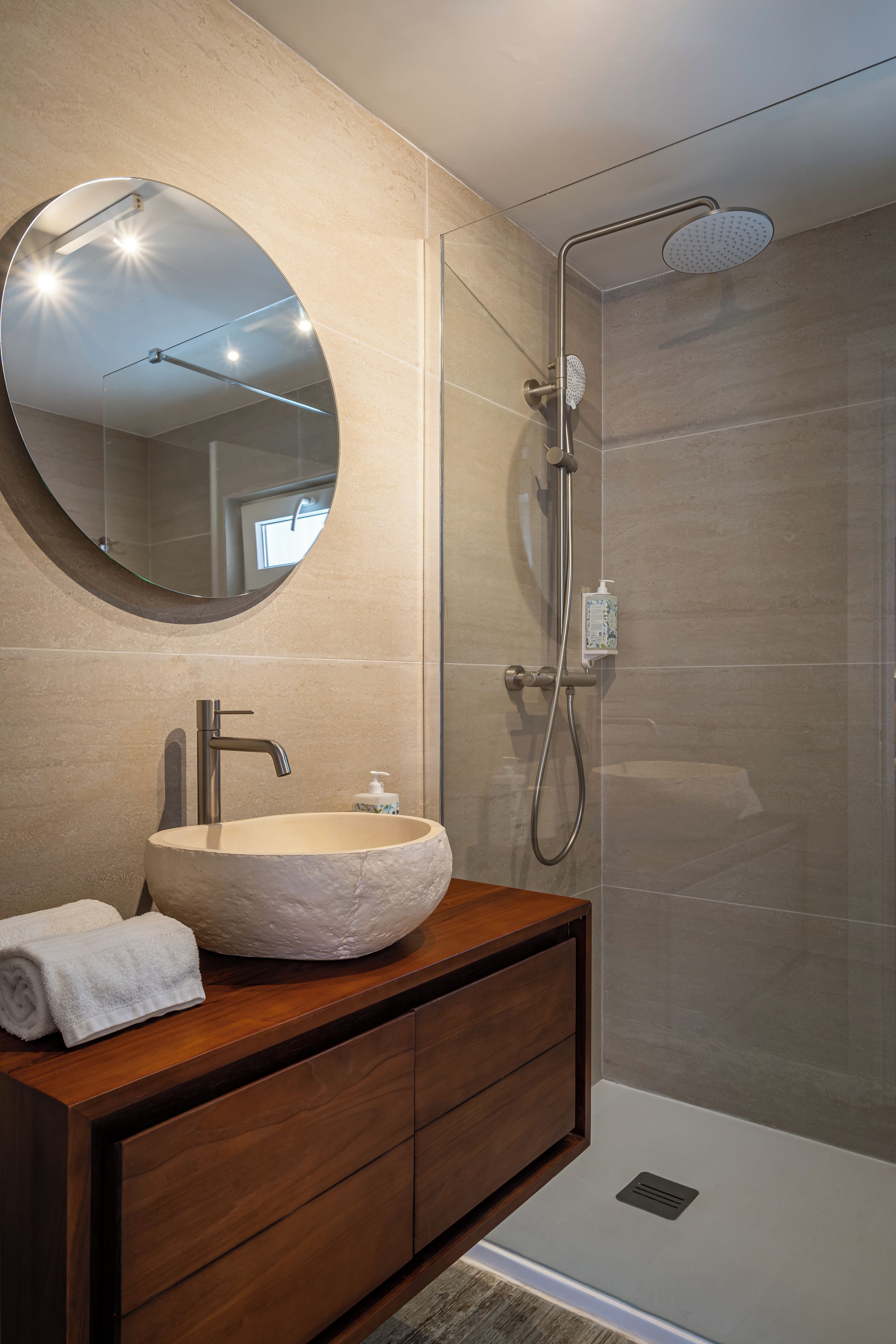 A bathroom with a round mirror, a vessel sink, a wooden vanity, and a glass-enclosed shower area.