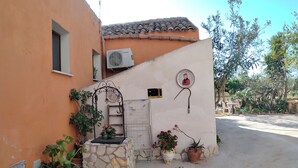 Un bâtiment avec un mur en pierre, une petite fenêtre et un portail en métal. On y trouve des plantes en pot et un banc en métal.