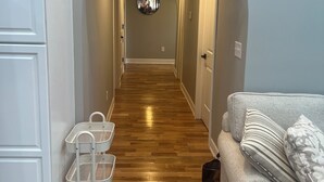 Un pasillo con suelo de madera, pared blanca y sofá gris con cojines estampados.