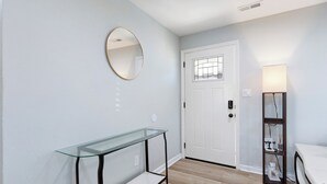 A modern room with a glass console table, a round mirror, a white door, and a floor lamp. - Charming 4-bedroom bungalow with AC and WiFi in Hampton, with washer and dryer. (Hampton)