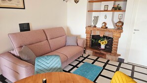 Un salon avec un canapé rose, un pouf bleu, une table basse en bois, une cheminée avec un manteau en briques et un tapis à motifs.