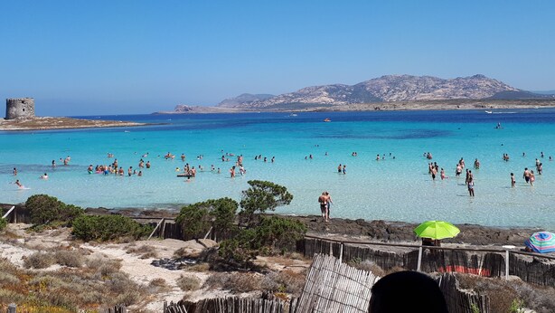 Une plage à l’eau turquoise et limpide, où l’on peut voir des gens nager et prendre des bains de soleil, avec une colline rocheuse en arrière-plan.