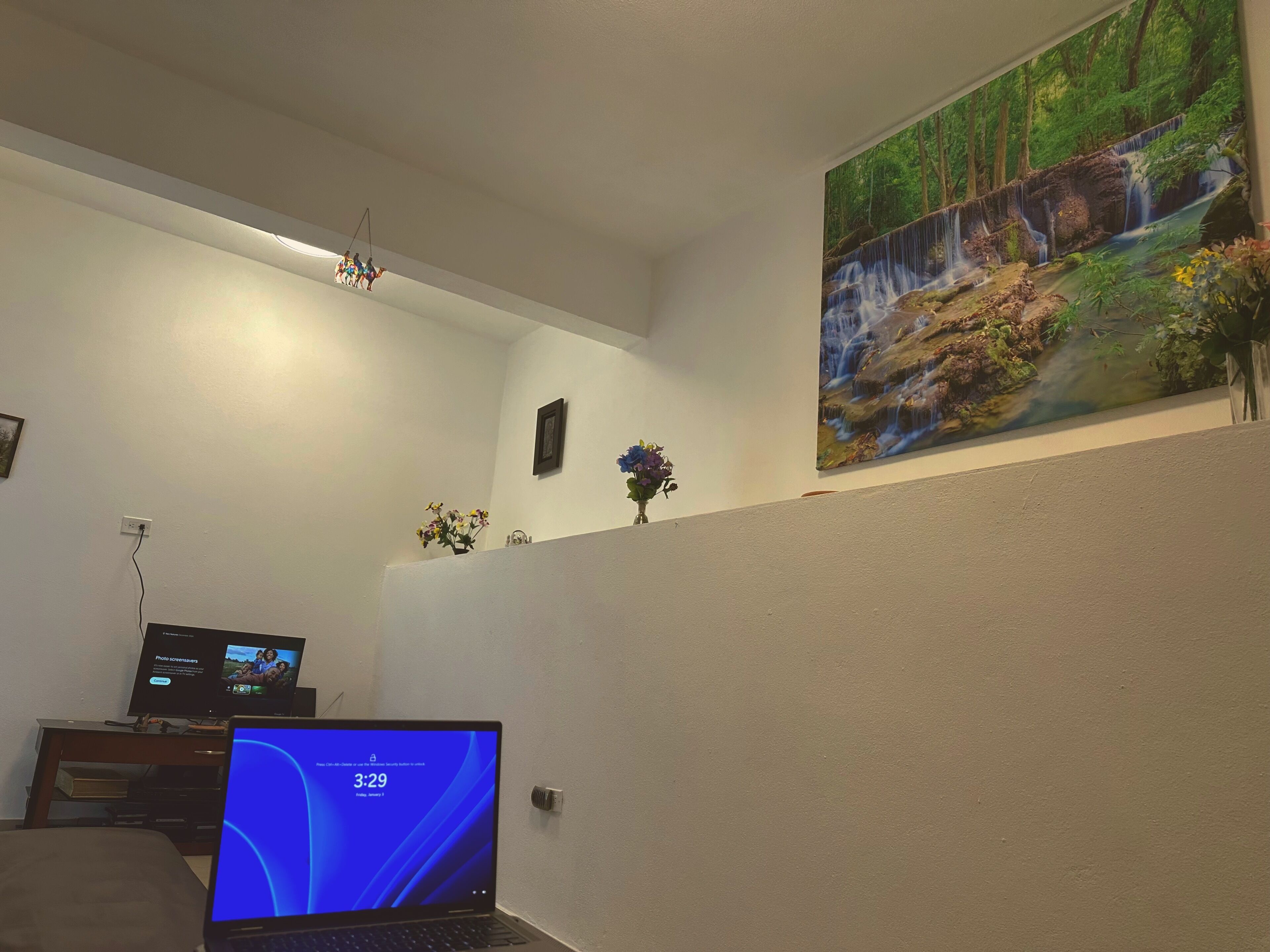Una habitación con una laptop sobre un escritorio, un televisor en un soporte, un cuadro colgado en la pared que muestra una cascada, y una lámpara en el techo.