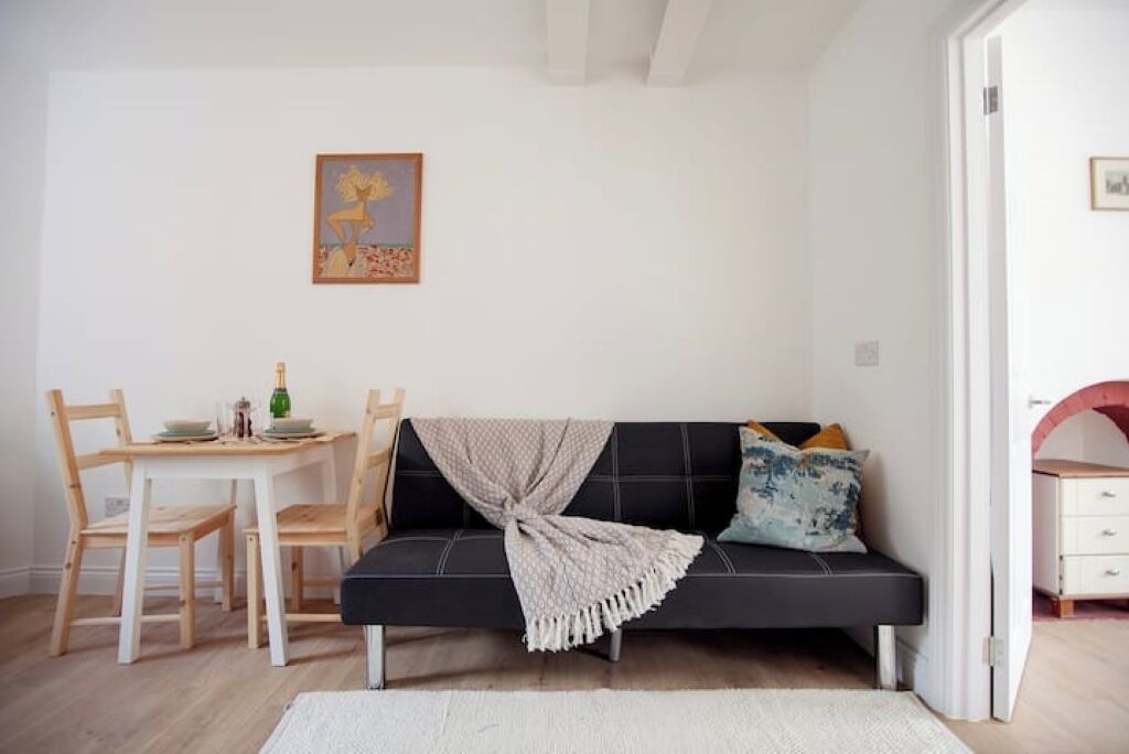 A room with a black sofa, wooden chairs, a small table with a bottle, and a painting on the wall.