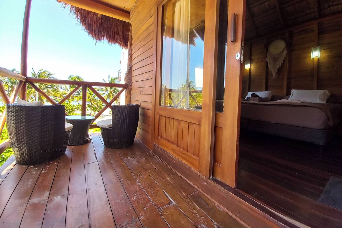 A wooden cabin with a balcony, a bed, wicker furniture, and a view of palm trees.