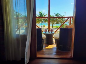 Vista desde una habitación con suelo de madera y muebles de mimbre, que da a una playa con palmeras y cielo despejado.