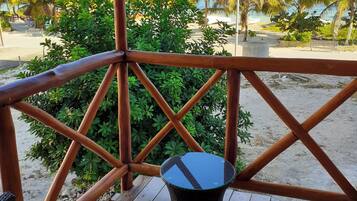 Um deck de madeira com móveis de vime e uma mesa, com vista para uma praia com palmeiras e céu claro.