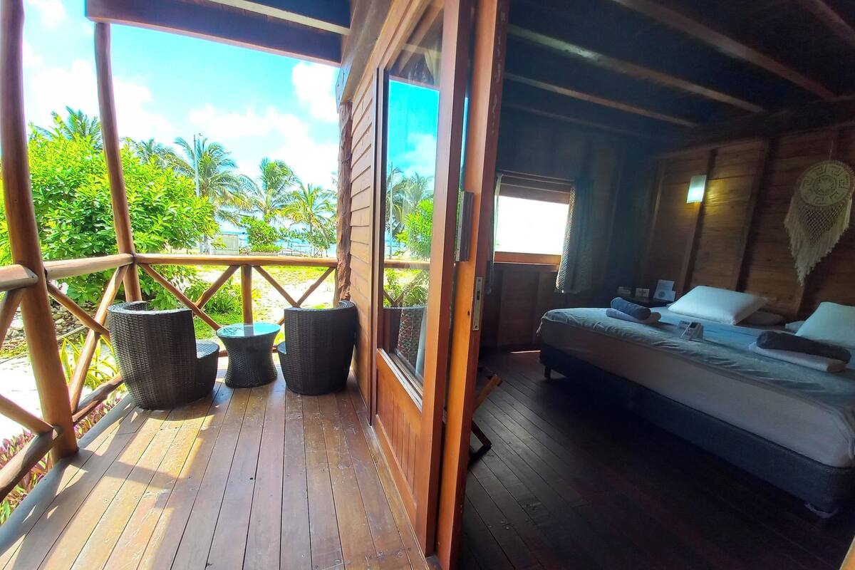 A wooden deck with wicker furniture, a bed with pillows and a remote control, and a view of palm trees.