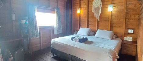A wooden cabin room with a thatched roof, a bed with white linens, a small table, and a wall-mounted light fixture.