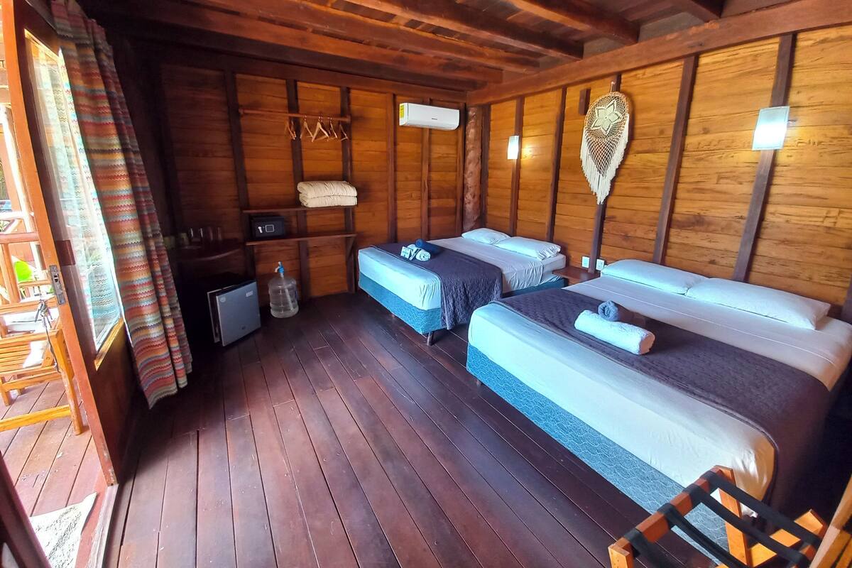 A wooden cabin room with two beds, a small table, a water dispenser, and a shelf with items.