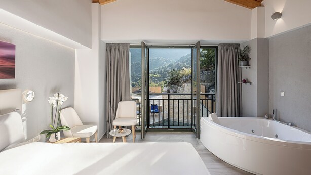 A modern bedroom with a large bed, a sitting area with a table and chairs, a bathtub, and a balcony with a scenic view.