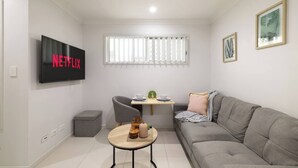 A modern living room with a grey sofa, a small round table with a decorative item, a grey armchair, a TV mounted on the wall, and a window with blinds. - Smart Suites Self Check-in Apartments - 35A Freney (Rocklea)