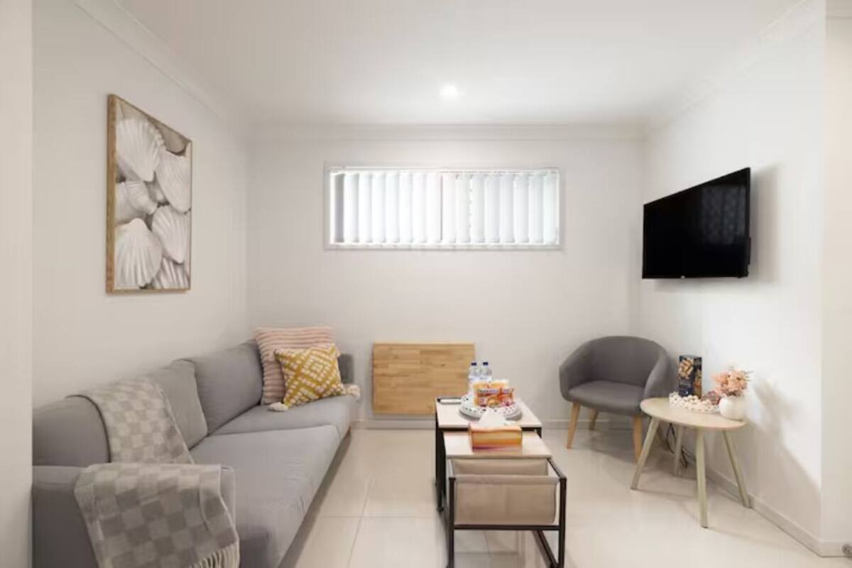 A modern living room with a grey sofa, a coffee table, a TV mounted on the wall, and a window with blinds.