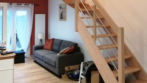 A modern living room with a wooden staircase, a grey sofa, and a small table. - Charming new 70 m² house in the heart of the Pines (L'Aiguillon-la-Presqu'île)