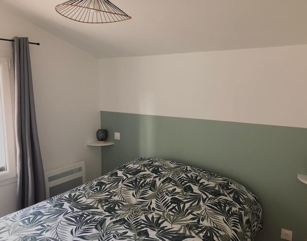 A bedroom with a bed covered in a tropical leaf-patterned bedspread, a grey curtain, a white radiator, and a small shelf with a decorative item.