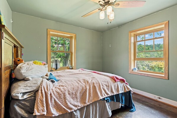 Un dormitorio con cama, dos ventanas, ventilador de techo y vistas al verdor exterior.